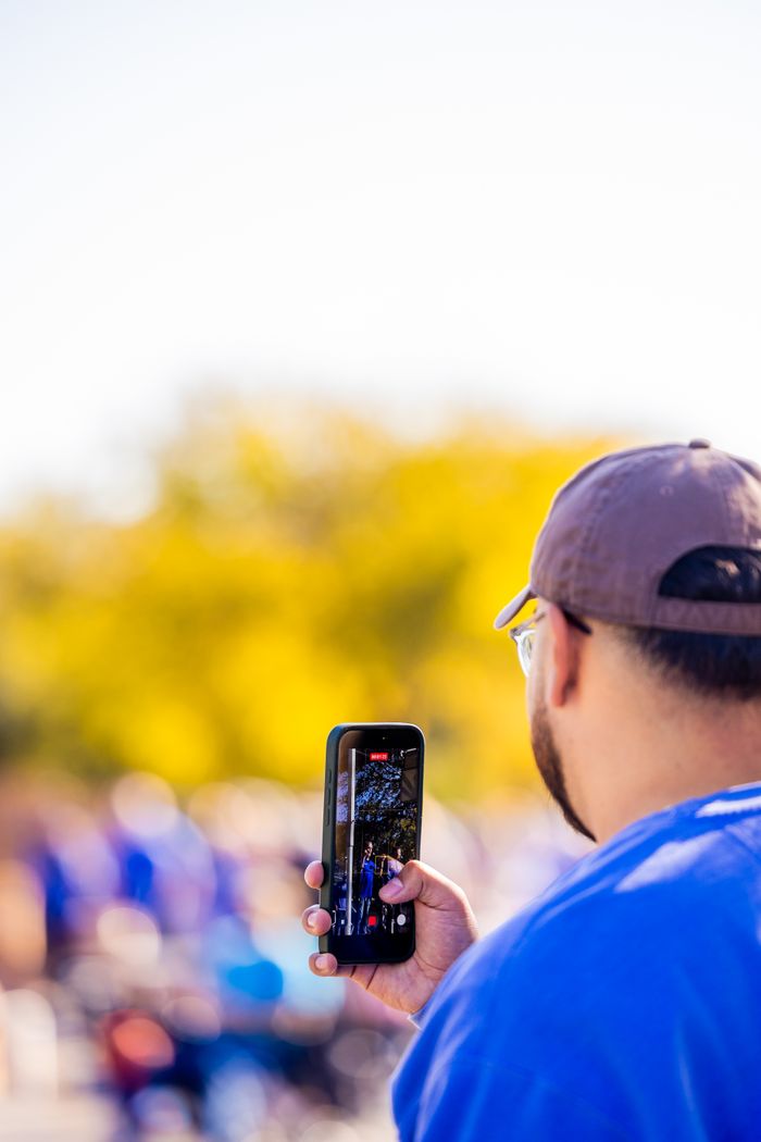 Adult capturing a lively outdoor event on a smartphone during a sunny day.